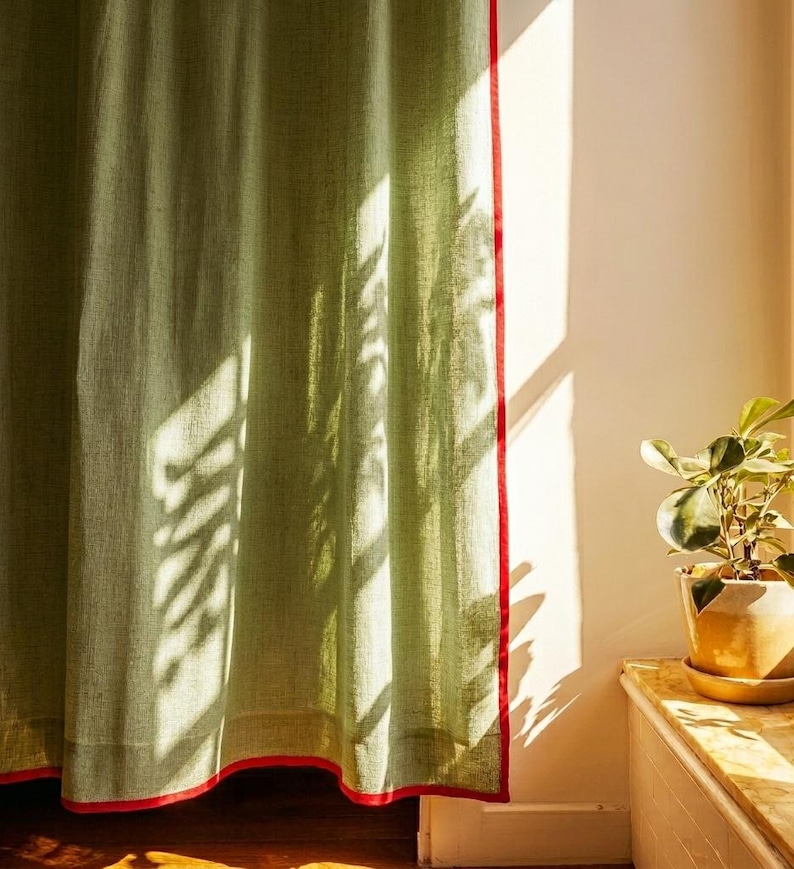 May include: A light green curtain with a red trim, casting shadows from a nearby plant. The curtain is made of a textured fabric and hangs in front of a white wall. A potted plant sits on a wooden surface.