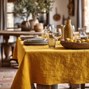 May include: A dining table set for a meal, featuring a mustard yellow linen tablecloth. The table is set with plates, glasses, and a wooden bowl filled with fruit. A tall, brown bottle and decorative vases are also present, creating a warm, inviting atmosphere.