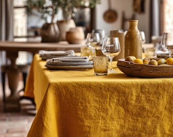 Könnte beinhalten: Ein Esstisch, der für eine Mahlzeit gedeckt ist, mit einer senfgelben Leinentischdecke. Der Tisch ist mit Tellern, Gläsern und einer Holzschale mit Obst gedeckt. Eine hohe, braune Flasche und dekorative Vasen sind ebenfalls vorhanden.
