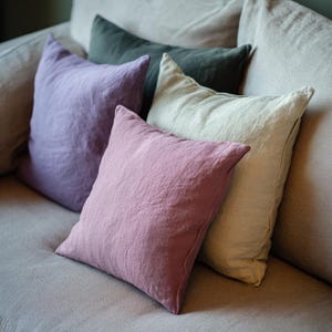 May include: A collection of square throw pillows in various colors, including lavender, dusty rose, sage green, and cream. The pillows are arranged on a light gray sofa, creating a cozy and inviting aesthetic.