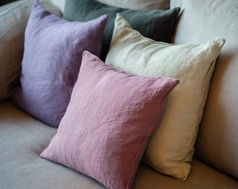 May include: A collection of square throw pillows in various colors, including lavender, dusty rose, sage green, and cream. The pillows are arranged on a light gray sofa, creating a cozy and inviting aesthetic.