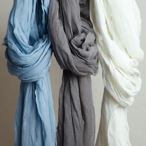 May include: Three linen scarves in shades of blue, gray, and white hang from a wooden rod. The scarves are draped, showcasing their soft texture and color variations. The background is a neutral, solid color.