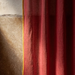 May include: A close-up of a red curtain with a yellow trim. The curtain is made of a textured fabric and hangs in soft folds. The background shows a textured wall with a shadow.