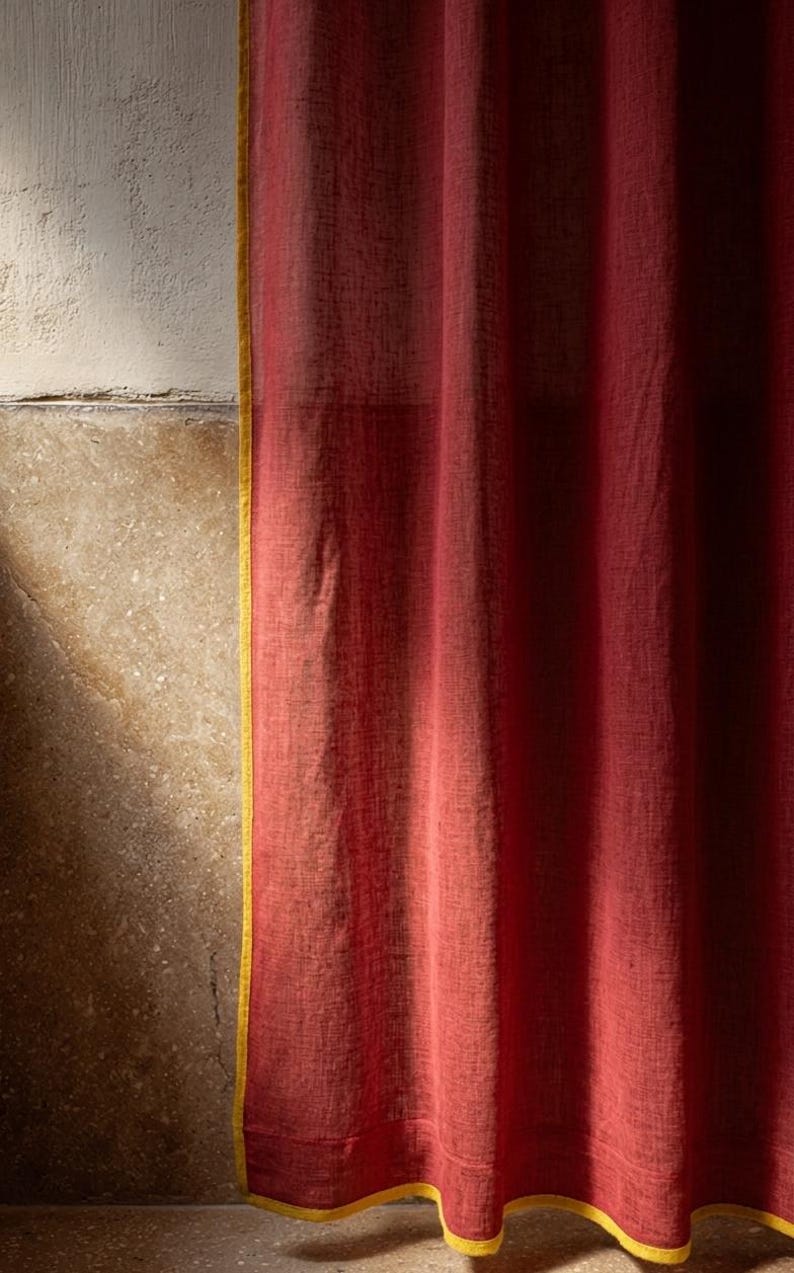 May include: A close-up of a red curtain with a yellow trim. The curtain is made of a textured fabric and hangs in soft folds. The background shows a textured wall with a shadow.