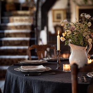 May include: A dining table set for a meal, featuring a dark tablecloth, plates, silverware, and a lit candle. A white pitcher with flowers and a staircase in the background complete the scene. The setting evokes a cozy, intimate atmosphere.