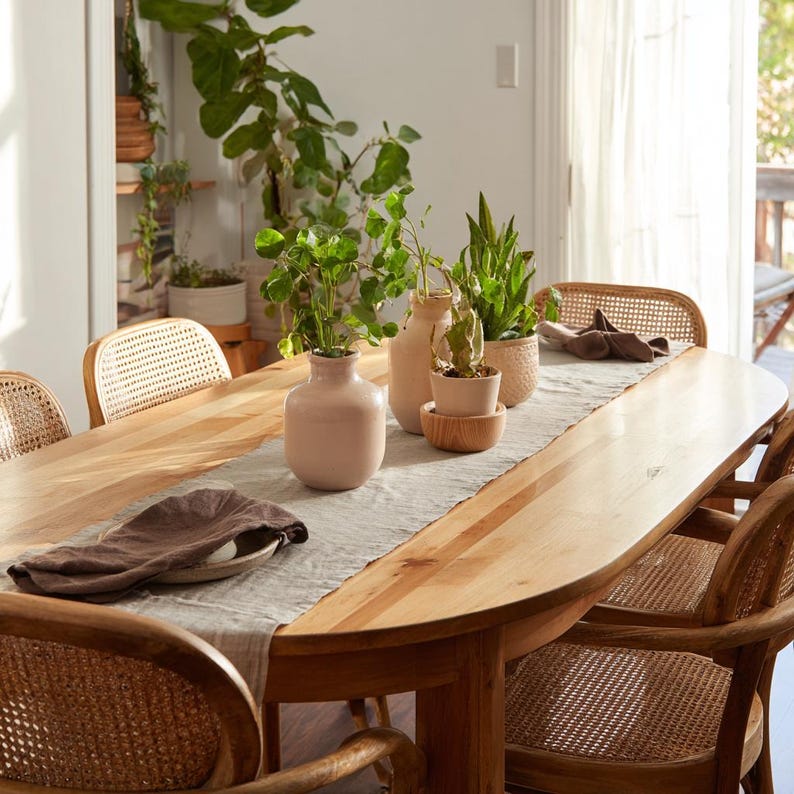 May include: A light wood dining table with a natural edge, set with a light gray runner and several decorative vases with plants. Wicker chairs surround the table, creating a warm and inviting dining space.