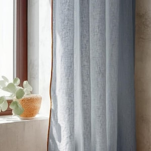 May include: Light blue linen curtains with a brown trim hang in front of a window. The curtains are partially drawn, allowing sunlight to filter through. A woven basket with a plant sits on the windowsill.