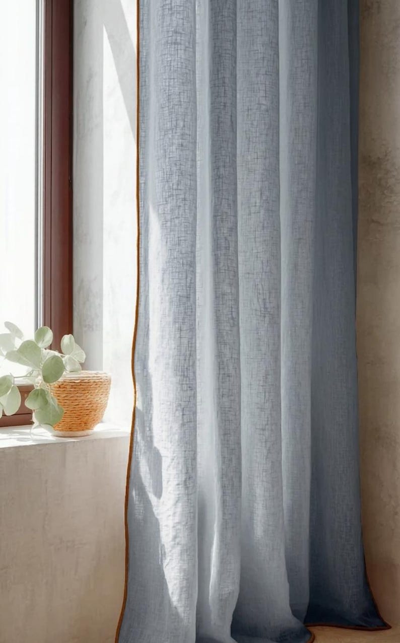May include: Light blue linen curtains with a brown trim hang in front of a window. The curtains are partially drawn, allowing sunlight to filter through. A woven basket with a plant sits on the windowsill.