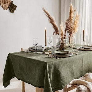 May include: A dining table set with a green linen tablecloth, plates, glasses, and cutlery. Tall candles and pampas grass in glass vases decorate the table. A neutral-toned throw blanket rests on a wooden chair.