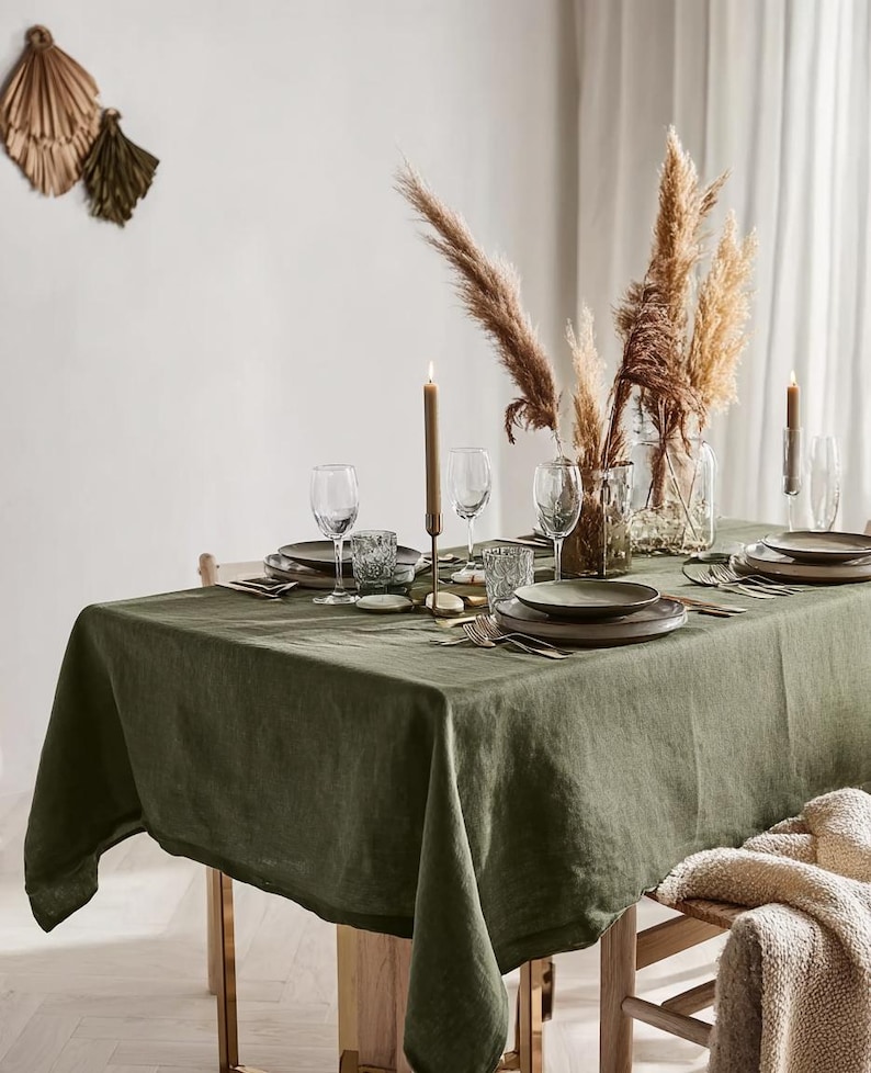 May include: A dining table set with a green linen tablecloth, plates, glasses, and cutlery. Tall candles and pampas grass in glass vases decorate the table. A neutral-toned throw blanket rests on a wooden chair.