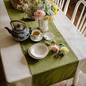May include: A table setting with a green runner, white tablecloth, and a floral arrangement. A teapot, teacup, and plates are arranged on the runner. The teapot is blue and white, and the teacup is filled with tea.