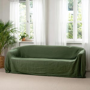 May include: A green sofa covered with a matching linen slipcover. The sofa is in front of a window with white sheer curtains. A large potted plant is to the left of the sofa.