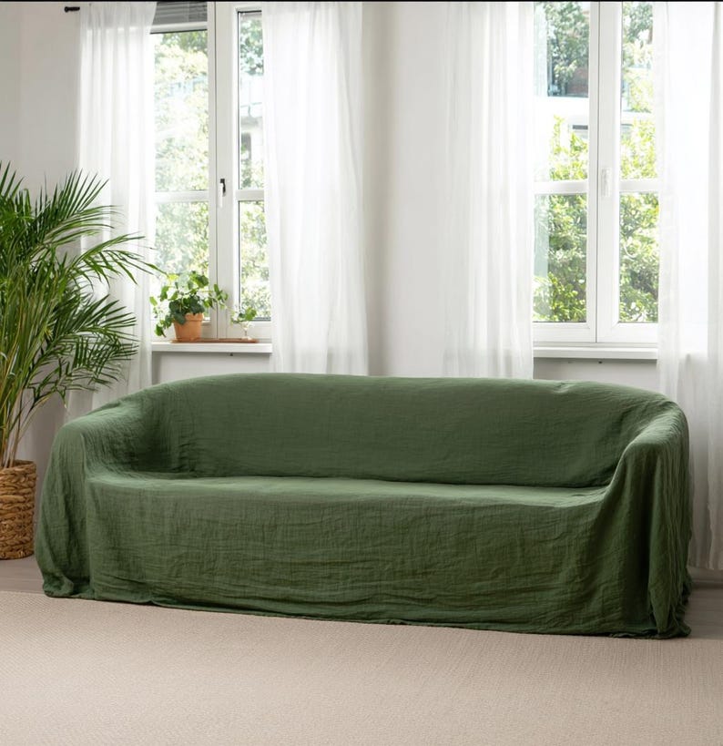 May include: A green sofa covered with a matching linen slipcover. The sofa is in front of a window with white sheer curtains. A large potted plant is to the left of the sofa.