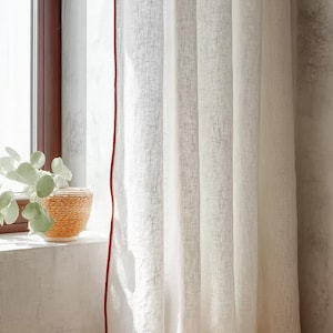 May include: White linen curtains with a red trim, hanging in front of a window. The curtains are slightly translucent, allowing light to filter through. A woven basket with a plant sits on the windowsill.