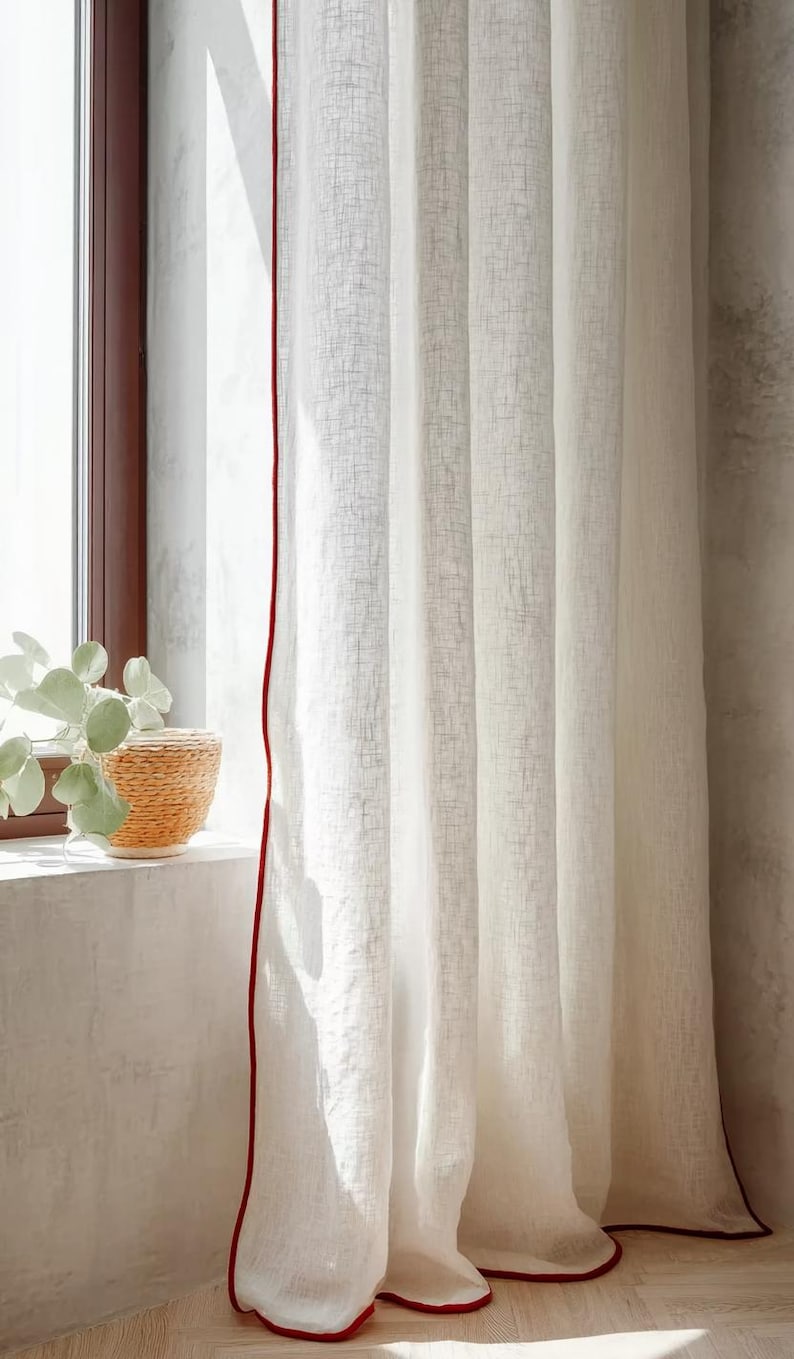 May include: White linen curtains with a red trim, hanging in front of a window. The curtains are slightly translucent, allowing light to filter through. A woven basket with a plant sits on the windowsill.