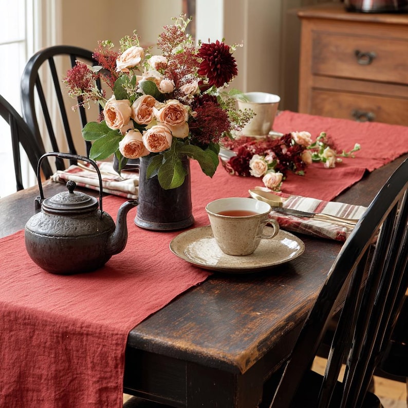 K&ouml;nnte beinhalten: Ein rustikaler Tisch f&uuml;r Tee. Eine dunkle Metallkanne steht auf einem roten Leinenl&auml;ufer, daneben eine Tasse und Untertasse. Eine Blumenvase und ein kleines Bouquet setzen Farbakzente. Schwarze St&uuml;hle umgeben den Tisch.