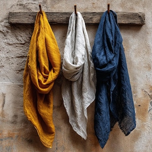 K&ouml;nnte beinhalten: Drei Leinen-Schals in Senfgelb, Hellgrau und Marineblau h&auml;ngen an einem Holzbalken vor einer strukturierten Wand. Die Schals sind drapiert und zeigen ihre weiche Textur und Farbvariationen. Die Wand hat ein rustikales, gealtertes Aussehen.