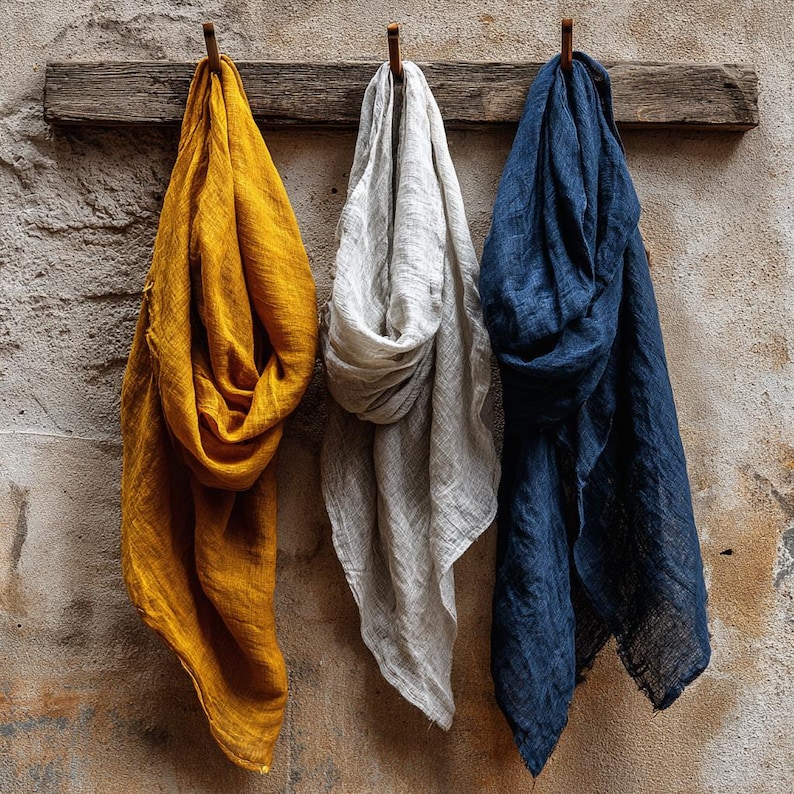 K&ouml;nnte beinhalten: Drei Leinen-Schals in Senfgelb, Hellgrau und Marineblau h&auml;ngen an einem Holzbalken vor einer strukturierten Wand. Die Schals sind drapiert und zeigen ihre weiche Textur und Farbvariationen. Die Wand hat ein rustikales, gealtertes Aussehen.