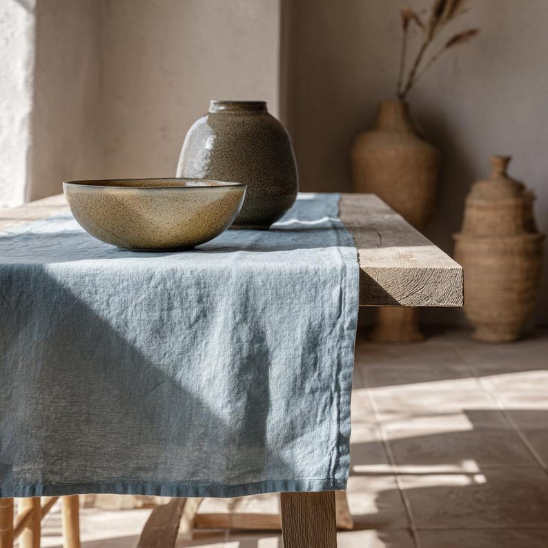 K&ouml;nnte beinhalten: Ein rustikaler Holztisch mit einem hellblauen Leinenl&auml;ufer. Eine gesprenkelte Keramikschale und eine strukturierte Vase stehen auf dem L&auml;ufer. Die Szene wird von nat&uuml;rlichem Licht durchflutet.