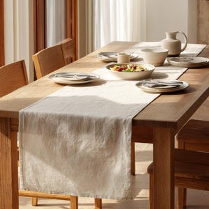 May include: A wooden dining table set for a meal. The table is made of light brown wood and has a cream-coloured table runner. The table is set with plates, bowls, silverware, and a jug. A salad is in a bowl.