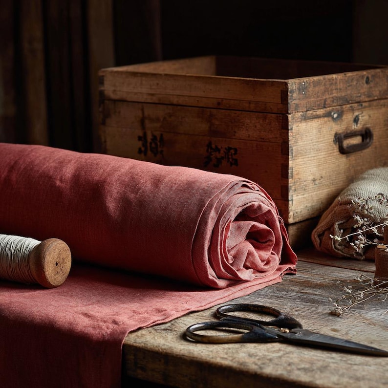 May include: A roll of terracotta-coloured fabric rests on a wooden table, with a wooden thread spool and vintage scissors. A wooden box is in the background, hinting at a craft or textile theme.
