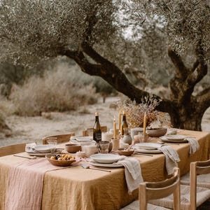 May include: Outdoor dining table set for a meal. The table is covered with a tan linen tablecloth and a pink runner. Dishes, bowls, glasses, and candles are arranged on the table. The scene is set outdoors under a large tree.