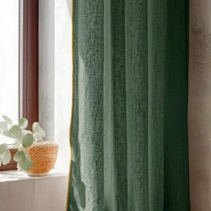May include: Green linen curtain with a textured weave and a gold trim. The curtain hangs in front of a window with a brown frame. A woven basket with a plant sits on the windowsill.