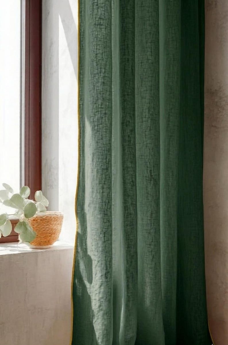 May include: Green linen curtain with a textured weave and a gold trim. The curtain hangs in front of a window with a brown frame. A woven basket with a plant sits on the windowsill.