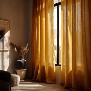 May include: Mustard yellow linen curtains hanging in front of a window, casting shadows on the floor. The curtains are long and flowing, with a textured appearance. A dark framed window is visible behind the curtains.