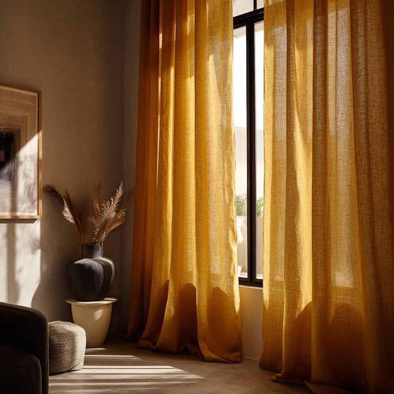 May include: Mustard yellow linen curtains hanging in front of a window, casting shadows on the floor. The curtains are long and flowing, with a textured appearance. A dark framed window is visible behind the curtains.
