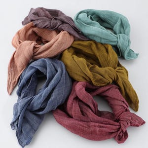 K&ouml;nnte beinhalten: Sieben geknotete Schals in verschiedenen Farben, darunter Staubrosa, T&uuml;rkis, Senfgelb, Tiefrot, Marineblau und ged&auml;mpftes Lila. Der Stoff scheint leicht und strukturiert zu sein, geeignet f&uuml;r w&auml;rmeres Wetter.