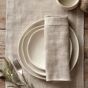 May include: A neutral-toned table setting featuring a linen table runner, white ceramic plates, a folded napkin, and a small bowl. Silverware and a sprig of greenery add a natural touch to the arrangement. The scene evokes a sense of rustic elegance.