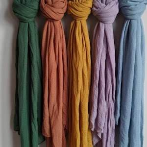 May include: Five colorful scarves hang from a wooden rod. The scarves are in shades of green, orange, yellow, purple, and blue. Each scarf is knotted at the top, with the fabric cascading downwards. The scarves are made of a soft, flowing material.