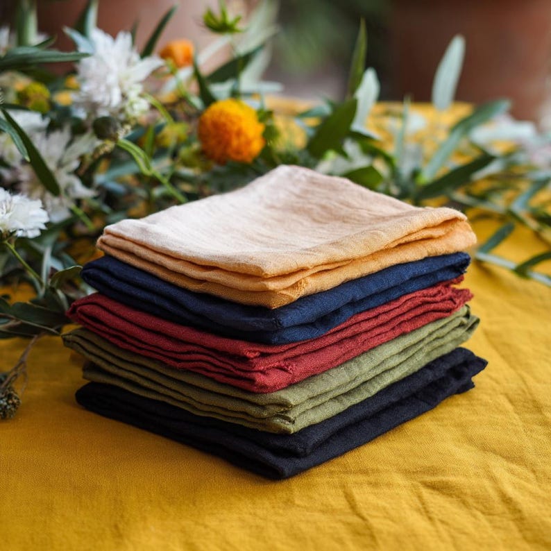 May include: A stack of seven folded linen napkins in colours including peach, navy, burgundy, olive green, and black. The napkins are on a yellow tablecloth, with flowers in the background.