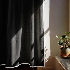 May include: A dark gray curtain with a white border hangs in front of a white wall. Sunlight streams through a window, casting shadows on the curtain. A potted plant sits on a wooden surface.