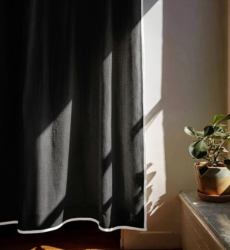 May include: A dark gray curtain with a white border hangs in front of a white wall. Sunlight streams through a window, casting shadows on the curtain. A potted plant sits on a wooden surface.