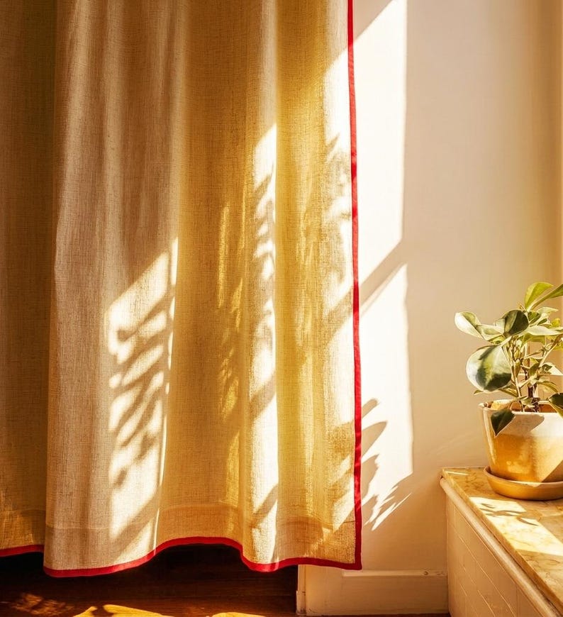 May include: Beige linen curtain with a vibrant red trim. The curtain is illuminated by sunlight, casting shadows. A potted plant sits on a marble ledge to the right. The curtain's texture is visible, adding depth to the image.