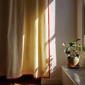 May include: Cream-colored linen curtain with a vibrant red trim. The curtain hangs in front of a white wall, with sunlight casting shadows. A potted plant sits on a windowsill to the right, adding a touch of greenery to the scene.