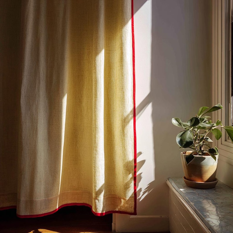 May include: Cream-colored linen curtain with a vibrant red trim. The curtain hangs in front of a white wall, with sunlight casting shadows. A potted plant sits on a windowsill to the right, adding a touch of greenery to the scene.
