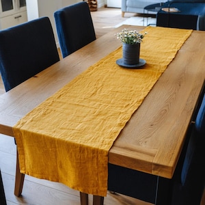 May include: A golden yellow linen table runner draped across a wooden dining table. A small vase with white flowers sits on a dark gray plate in the center. Dark blue chairs surround the table.