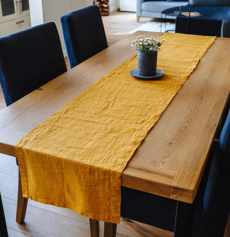 May include: A golden yellow linen table runner draped across a wooden dining table. A small vase with white flowers sits on a dark gray plate in the center. Dark blue chairs surround the table.