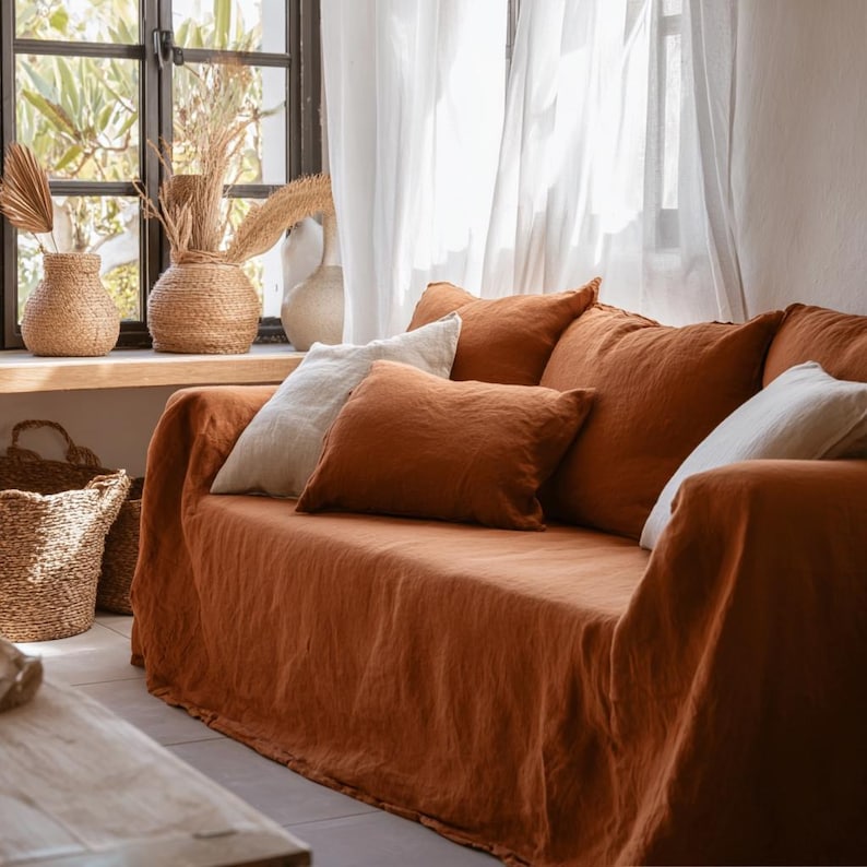 May include: A living room scene featuring a rust-colored sofa draped with a matching linen cover. Several throw pillows in white and rust hues are arranged on the sofa. Woven vases and baskets add a natural touch, complemented by sheer white curtains and a window.