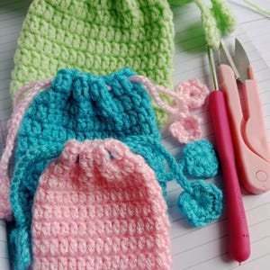 May include: A collection of handmade crochet drawstring bags in various sizes and colors. The bags are in shades of green, teal, and pink, with matching drawstrings. A crochet hook and small scissors are also visible, suggesting a crafting theme.