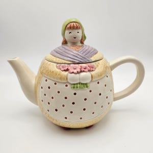 May include: A ceramic teapot with a whimsical design. The teapot features a woman's head as the lid, wearing a green bonnet and a purple shawl. The body is cream-colored with brown polka dots and a floral bouquet.