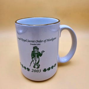 May include: White ceramic mug with a gold rim and a curved handle. The mug features green text and a graphic of a leprechaun with a walking stick. The text reads "Loyal Royal Secret Order of Hooligans, Founded 1982" and "2003".