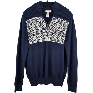 May include: Navy blue zip-up sweater with a white geometric pattern across the chest. The sweater has a ribbed collar and cuffs, and a small logo tag near the zipper. The sweater is hanging on a black hanger.