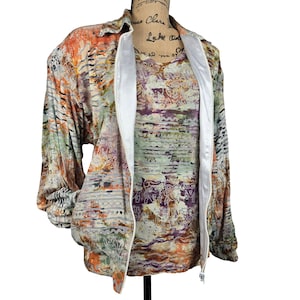 May include: A zip-up jacket featuring a multi-colored abstract pattern in shades of orange, purple, green, and white. The jacket has a white zipper placket, long sleeves with elastic cuffs, a collar, and a matching patterned shirt underneath.