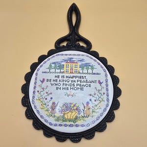 May include: A black cast iron trivet with a decorative handle and a round, scalloped frame. The center features a cross-stitch design with the text "HE IS HAPPIEST, BE HE KING OR PEASANT WHO FINDS PEACE IN HIS HOME."