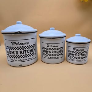 May include: A set of three white enamel canisters with lids, each featuring a black checkered pattern and the text "Welcome MOM'S KITCHEN We're always open" printed on the front. The canisters vary in size, decreasing from left to right.