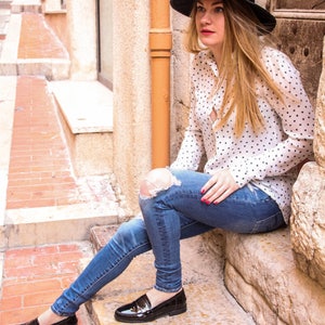 May include: A woman wearing a white polka dot shirt, blue jeans with a distressed knee, and black loafers sits on stone steps. She is wearing a black hat with a black ribbon.